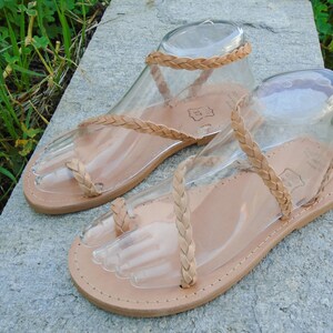 May include: A pair of tan leather sandals with braided straps. The sandals have a toe loop and an ankle strap.
