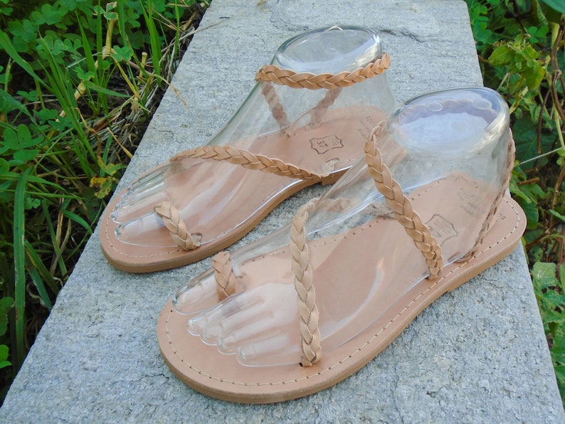 May include: A pair of tan leather sandals with braided straps. The sandals have a toe loop and an ankle strap.