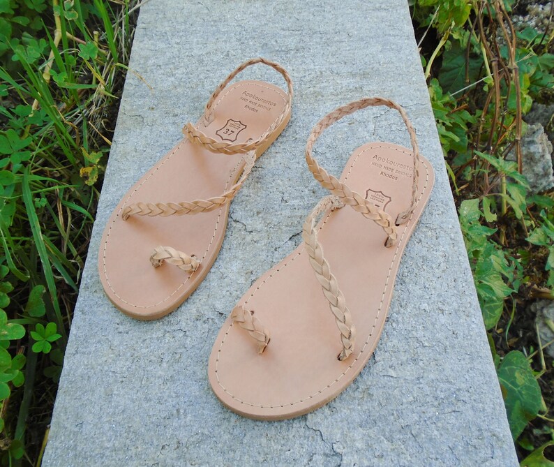 May include: A pair of tan leather sandals with braided straps. The sandals have a toe loop and an ankle strap. The sandals are made by "Andalousite Made with Pride Athens".