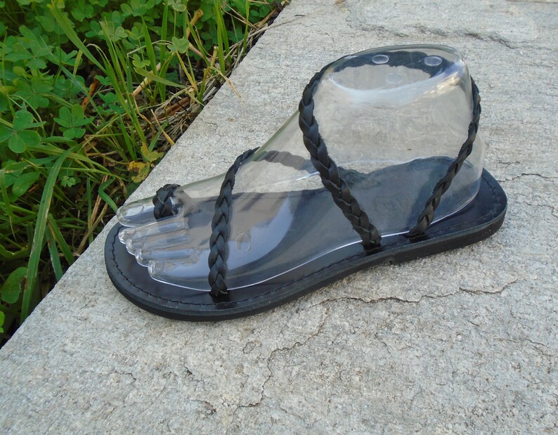 May include: Black leather sandals with braided straps. The sandals have a flat sole and are designed to be worn with bare feet.