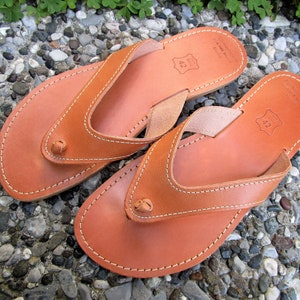 May include: A pair of brown leather thong sandals with a simple design. The sandals have a white stitch detail around the edges and a small leather strap across the toe. The sandals are size 42.