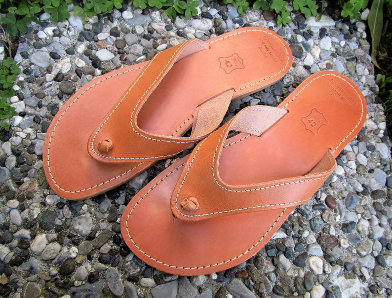 May include: A pair of brown leather thong sandals with a simple design. The sandals have a white stitch detail around the edges and a small leather strap across the toe. The sandals are size 42.