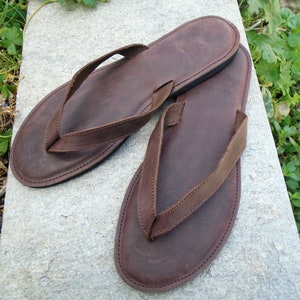 May include: A pair of brown leather flip-flops. The sandals have a dark brown sole and a matching thong strap. The flip-flops are placed on a gray stone surface, with green foliage in the background. The leather appears to be distressed.