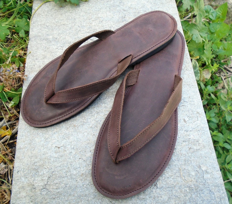 May include: A pair of brown leather flip-flops. The sandals have a dark brown sole and a matching thong strap. The flip-flops are placed on a gray stone surface, with green foliage in the background. The leather appears to be distressed.