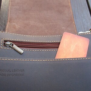 May include: Close-up of a brown leather bag with a zippered pocket and a brown card inside. The bag has a label that reads "ORIGINAL LEATHER MADE IN GREECE".