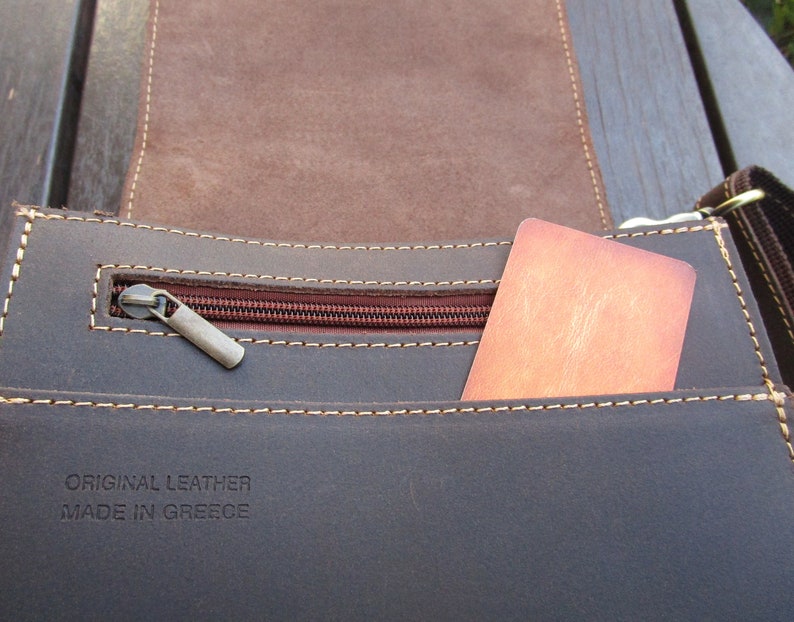 May include: Close-up of a brown leather bag with a zippered pocket and a brown card inside. The bag has a label that reads "ORIGINAL LEATHER MADE IN GREECE".