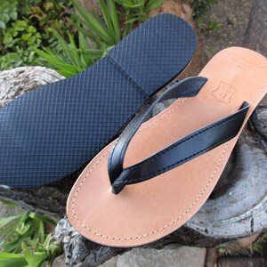 May include: A pair of leather flip-flops with a black strap and a light brown footbed. The sole is dark blue. The flip-flops are displayed on a natural wood surface, suggesting a handcrafted or artisanal product.