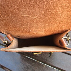May include: The inside of a brown leather bag with a flap closure. The bag has a brown leather strap and a metal buckle. The bag is empty.