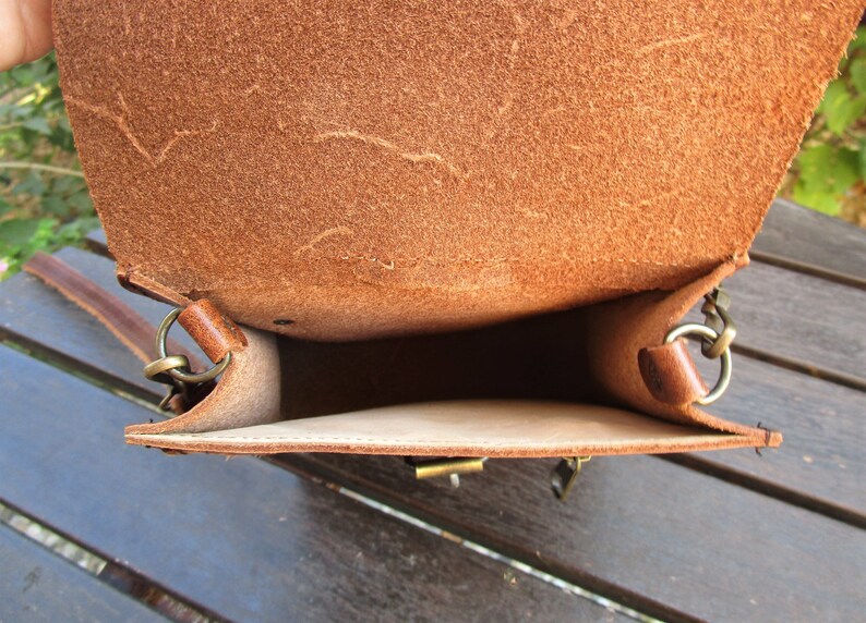 May include: The inside of a brown leather bag with a flap closure. The bag has a brown leather strap and a metal buckle. The bag is empty.