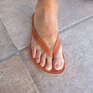 May include: A brown leather flip-flop with white stitching. The sandal has a simple thong design and a rounded toe. The image shows the sandal on a foot, resting on a stone surface.
