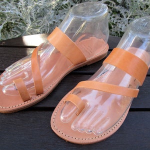 May include: A pair of light brown leather sandals with a toe loop and ankle strap. The sandals are displayed on clear plastic foot forms. The soles are stitched with a contrasting thread. The sandals are on a dark wooden surface.