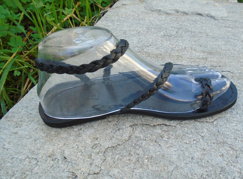 May include: Black leather sandals with braided straps. The sandals have a flat sole and are made of a soft, supple leather.