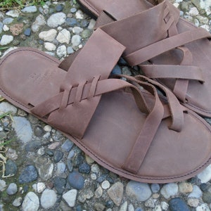 May include: A pair of brown leather sandals with multiple straps. The sandals have a braided design on the straps and a simple, rounded toe.
