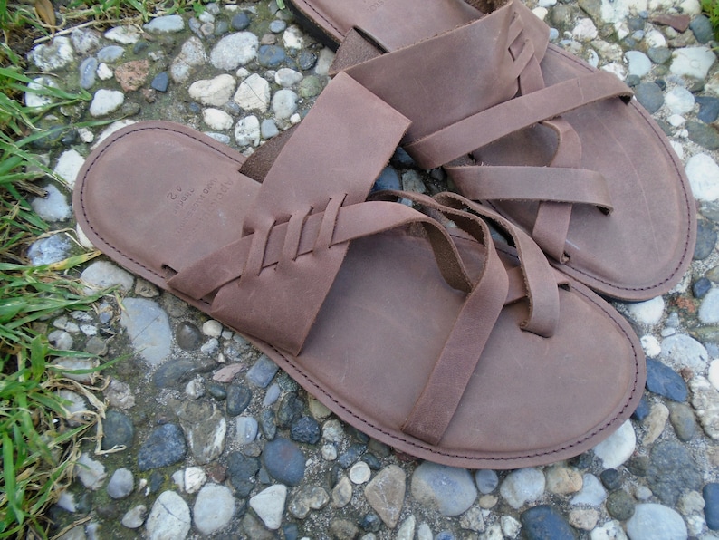 May include: A pair of brown leather sandals with multiple straps. The sandals have a braided design on the straps and a simple, rounded toe.
