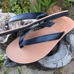 May include: A pair of black leather flip-flops with a light brown footbed. The straps are black and stitched to the footbed. The flip-flops are displayed on a weathered wooden surface.