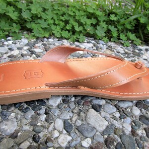 May include: Tan leather thong sandals with a simple design. The sandals have a leather strap that goes between the toes and a leather strap that goes around the ankle. The sandals are made of high-quality leather and are perfect for summer.