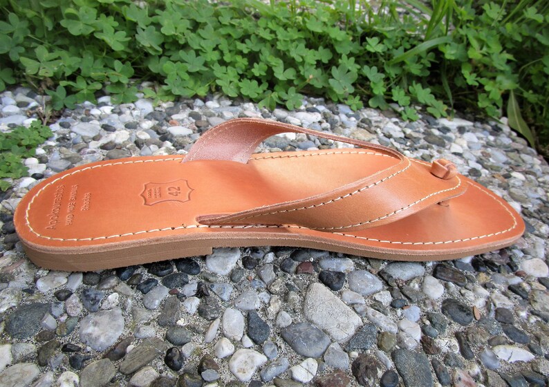 May include: Tan leather thong sandals with a simple design. The sandals have a leather strap that goes between the toes and a leather strap that goes around the ankle. The sandals are made of high-quality leather and are perfect for summer.