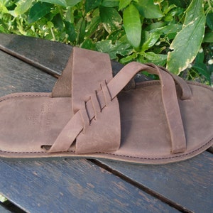 May include: Brown leather sandals with a braided strap design. The sandals have a simple, minimalist style.