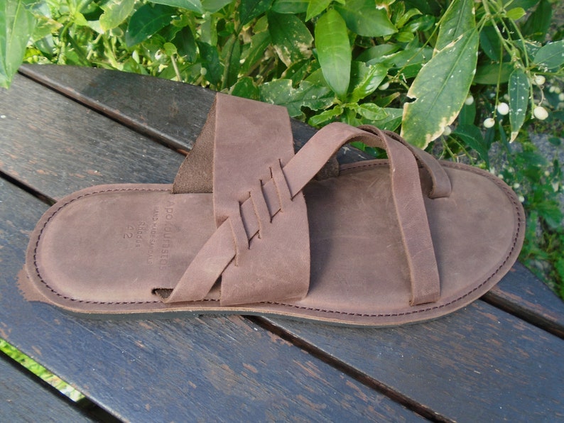 May include: Brown leather sandals with a braided strap design. The sandals have a simple, minimalist style.