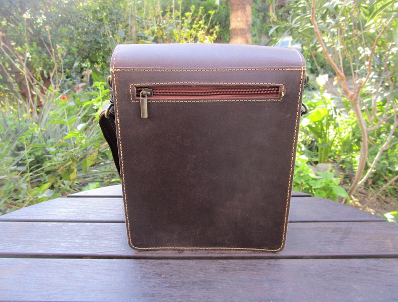 May include: A brown leather messenger bag with a zippered pocket on the back. The bag has a flap closure and a strap.