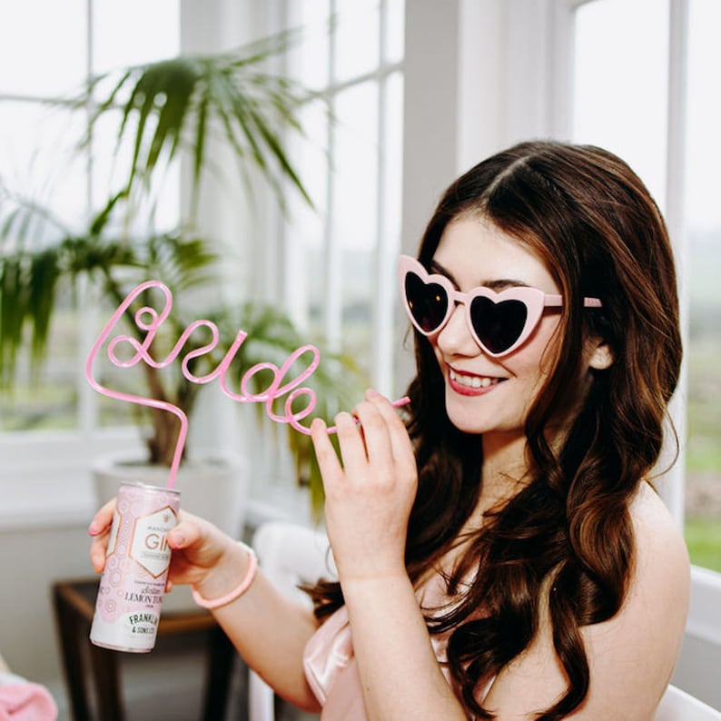 Image shows a  dark haired woman wearing pink heart shaped sunglasses drinking from a can using a pink straw in the shape of the word "Bride"