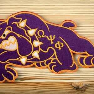 May include: Purple embroidered patch of a bulldog with white teeth and orange outline. The bulldog has a spiked collar with white spikes. The letters "ΩΨΦ" are embroidered in yellow on the bulldog's back.