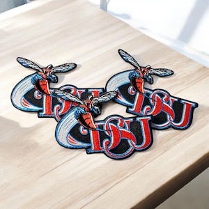 May include: Three embroidered patches with a black background and the letters "CSU" in red with a white outline. Each patch features a stylized red and blue insect design above the letters. The patches are on a light wood surface.