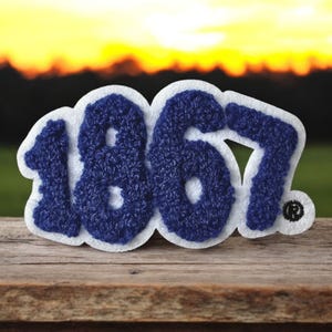May include: A blue and white felt patch with the number "1867" stitched in a textured, dark blue thread. The patch is on a wooden surface with a blurred sunset background.