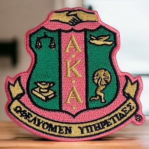 Könnte beinhalten: Rosa-grüner gestickter Aufnäher mit einem Wappen, das eine Justizwage, ein Buch, einen Vogel und eine Figur mit einem Globus zeigt. Das Wappen ist von einem goldenen Band mit dem Text "AKA" und "WOMEN OF YESTERDAY" umgeben.