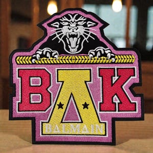 Può includere: Toppa ricamata con sfondo rosa, con le lettere "BAK" in rosso e giallo, e la parola "BALMAIN" in bianco. Una pantera nera con dettagli bianchi si trova sopra le lettere. La toppa ha un bordo nero.