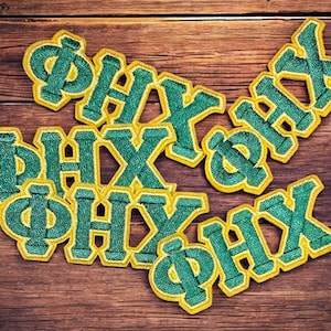 Pode incluir: Remendos bordados com as letras gregas ΦΗΧ em verde com borda amarela. Os remendos são dispostos em uma superfície de madeira, criando uma exibição visualmente atraente. Estes remendos são ideais para personalizar roupas ou acessórios.