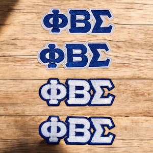 May include: Four embroidered patches with the Greek letters ΦΒΣ in a bold, blocky font. The letters are in a deep blue color with a white border. The patches are arranged on a light brown wooden surface.