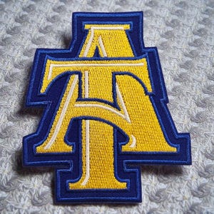 May include: Embroidered patch featuring the letters "AT" in a bold, stylized design. The letters are yellow with white accents, outlined in a deep blue border. The patch is likely for apparel or accessories.