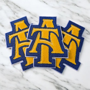 May include: Three blue and gold varsity letter patches. Each patch features the letters "AT" in gold, outlined in blue. The patches are arranged on a white marble surface.