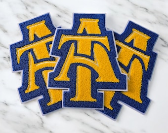 May include: Three blue and gold varsity letter patches. Each patch features the letters "AT" in gold, outlined in blue. The patches are arranged on a white marble surface.