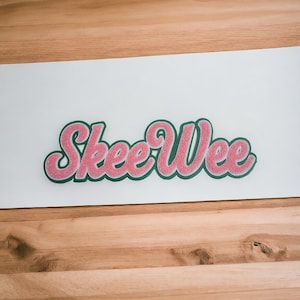May include: Pink and green felt letters spelling out the word "Skee Wee" on a white surface.