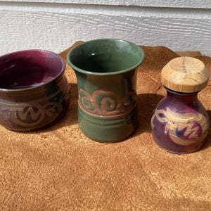 May include: Three handcrafted ceramic pieces: a small bowl with a dark purple interior and brown exterior with a decorative pattern, a green vase with a reddish-brown design, and a small jar with a wooden lid, featuring purple and gold accents.