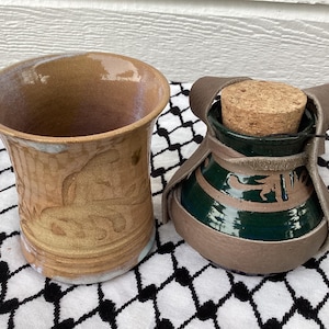 May include: Two ceramic vessels. One is a tall, open-topped mug with a light brown glaze and etched designs. The other is a green and brown ceramic flask with a cork stopper, encased in a brown leather wrap.
