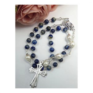May include: A rosary featuring dark blue mottled beads and white pearl-like beads connected by silver-toned metal links. The piece includes a silver-toned metal crucifix at the bottom and a small silver-toned medal depicting a religious figure.