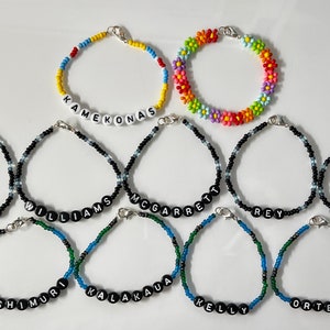 May include: A collection of 11 beaded bracelets with different names spelled out in black beads. The bracelets are made with black, white, blue, green, yellow, and red beads. Some bracelets have a flower pattern.