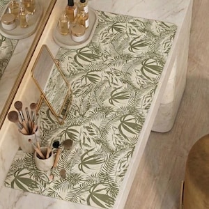 May include: A vanity table with a marble top features a green and white tropical leaf print mat. Makeup brushes, mirrors, and bottles of beauty products are arranged on the surface, creating a stylish and functional space.