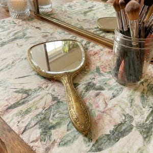 May include: A gold-toned hand mirror with an ornate handle rests on a floral patterned surface. A rectangular mirror and a jar of makeup brushes are in the background. The scene evokes a vintage vanity setting.
