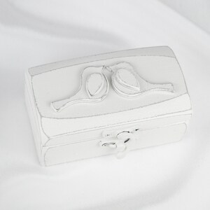 May include: White wooden ring box with a carved design of two birds facing each other. The box has a hinged lid and a small metal latch.