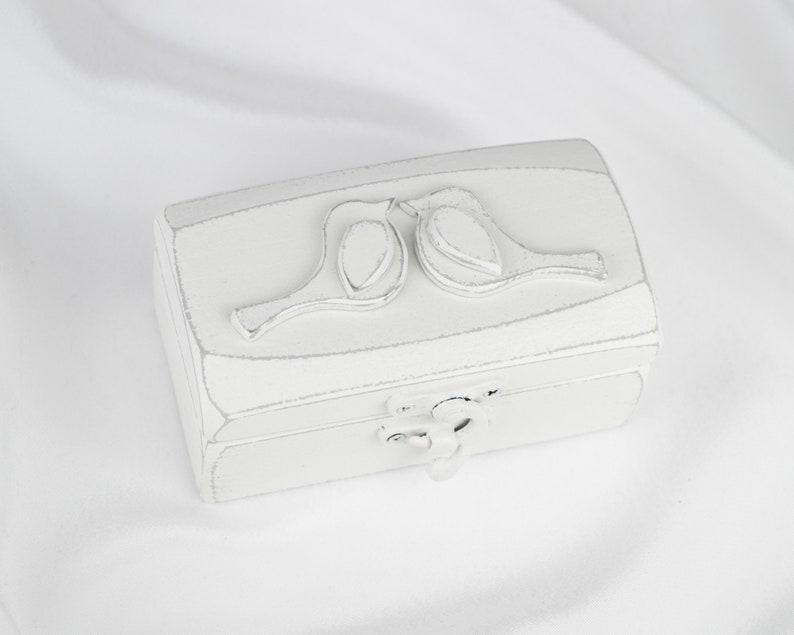 May include: White wooden ring box with a carved design of two birds facing each other. The box has a hinged lid and a small metal latch.