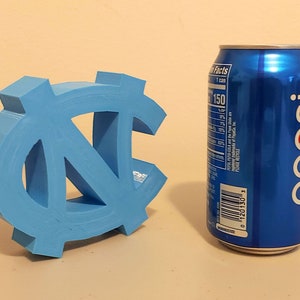 May include: A light blue 3D printed UNC Tar Heels logo next to a blue Pepsi can. The can has nutrition facts and a barcode on the side.