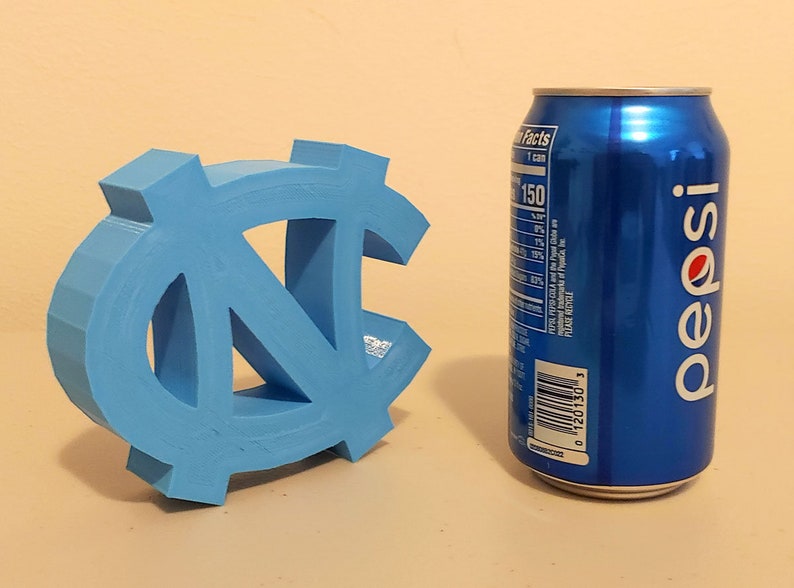 May include: A light blue 3D printed UNC Tar Heels logo next to a blue Pepsi can. The can has nutrition facts and a barcode on the side.