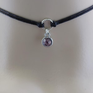 May include: A black choker necklace with a silver-colored ring and a small, round, light purple gemstone pendant. The necklace is made of two strands of black cord.
