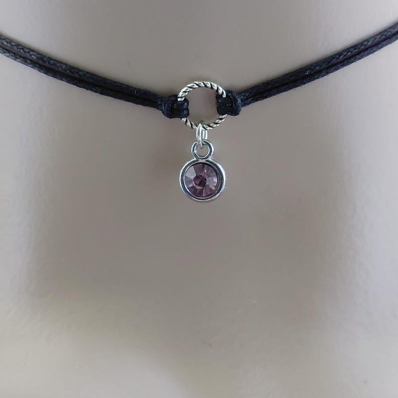 May include: A black choker necklace with a silver-colored ring and a small, round, light purple gemstone pendant. The necklace is made of two strands of black cord.