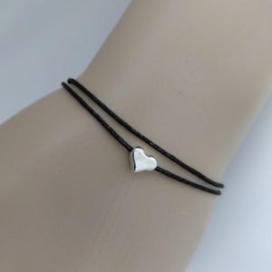 May include: A double-strand black cord bracelet featuring a single, small, polished silver-tone heart charm. The bracelet is constructed from two thin, dark, braided cords that wrap around the wrist, with the metallic heart centred on one strand.