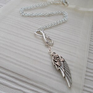Pu&ograve; includere: Collana color argento con un simbolo dell'infinito e un ciondolo a forma di ala con una rosa. La collana &egrave; esposta su un tessuto color crema, evidenziando il suo design delicato e i dettagli intricati.